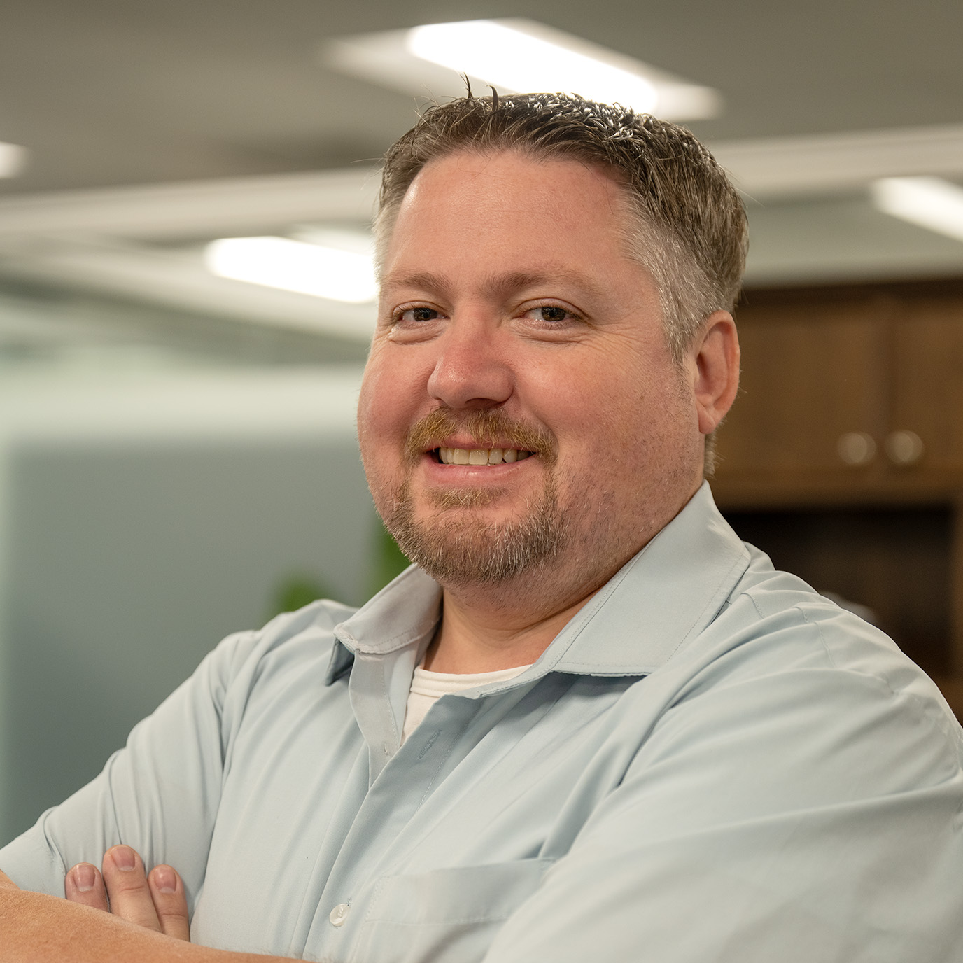 Man with short hair and goatee smiling, wearing a light blue shirt, indoors with arms crossed.