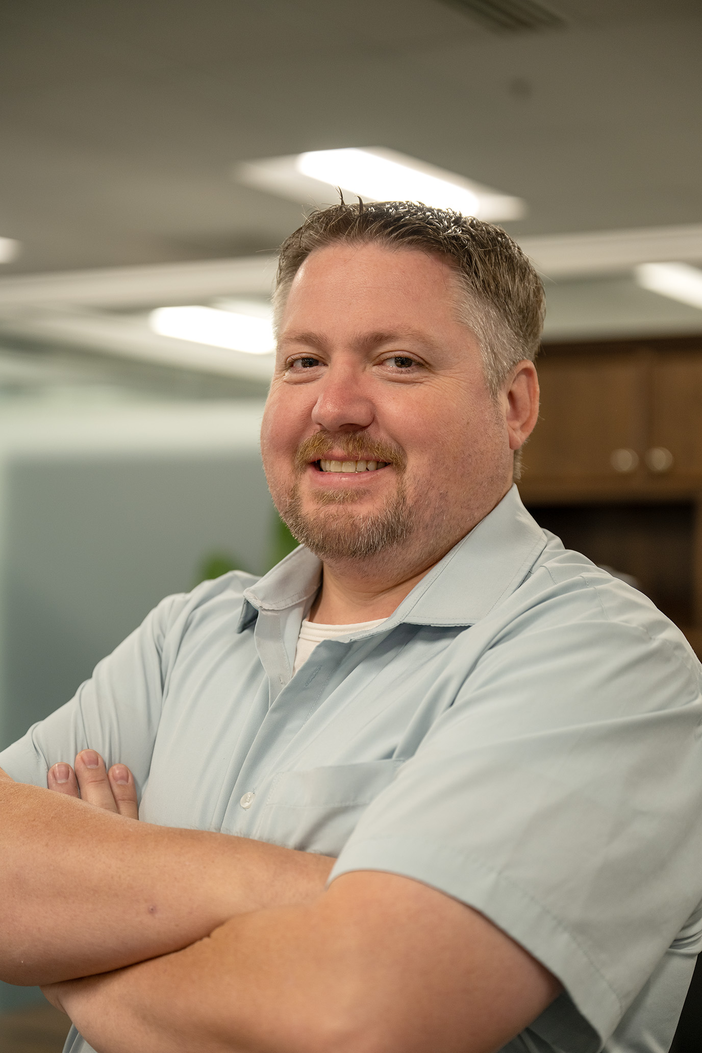 Man with short hair and a beard smiling, arms crossed, wearing a light blue shirt indoors.